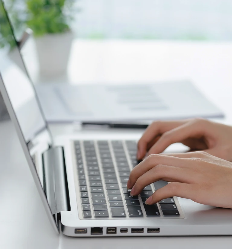 Close-up of hands typing on a silver laptop keyboard with a blurred document and plant in the background.