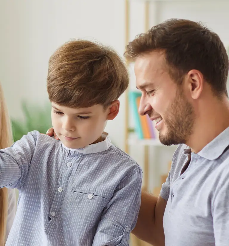 A man smiling and gently touching a young boy wearing a light blue striped shirt indoors.