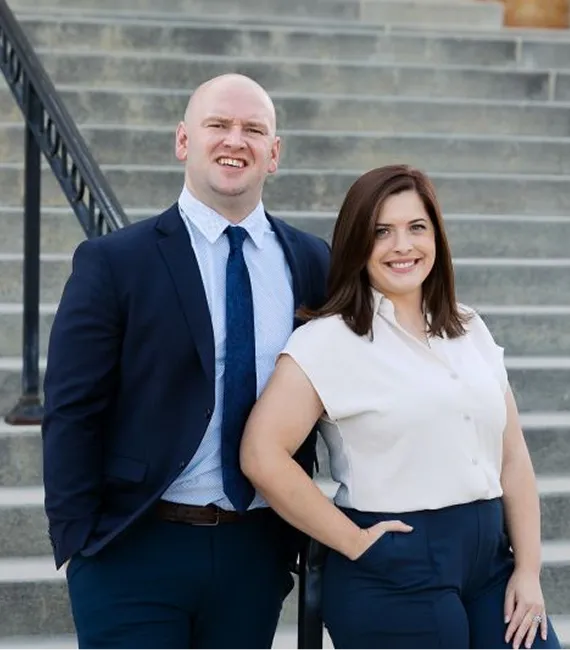 Two professionally dressed individuals, a man in a navy suit and a woman in a white blouse and navy pants, standing on outdoor stone steps.