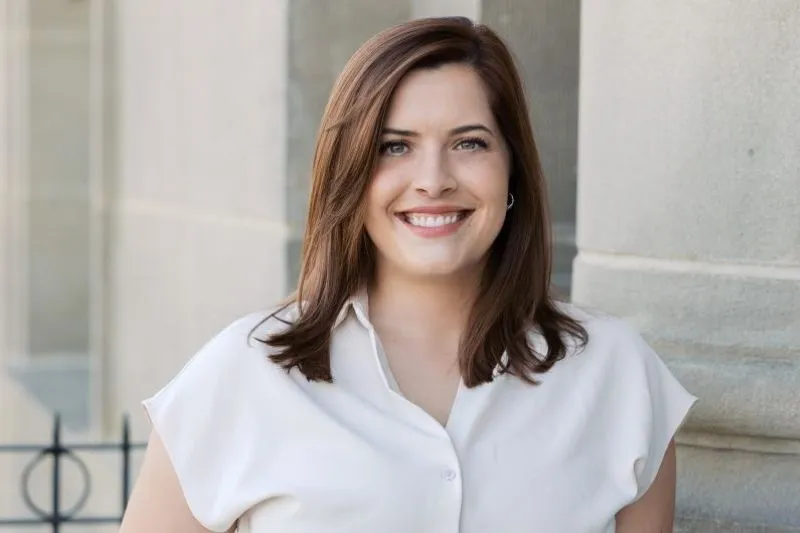 Portrait of a smiling woman with shoulder-length brown hair, wearing a white blouse, standing outdoors near a stone wall.