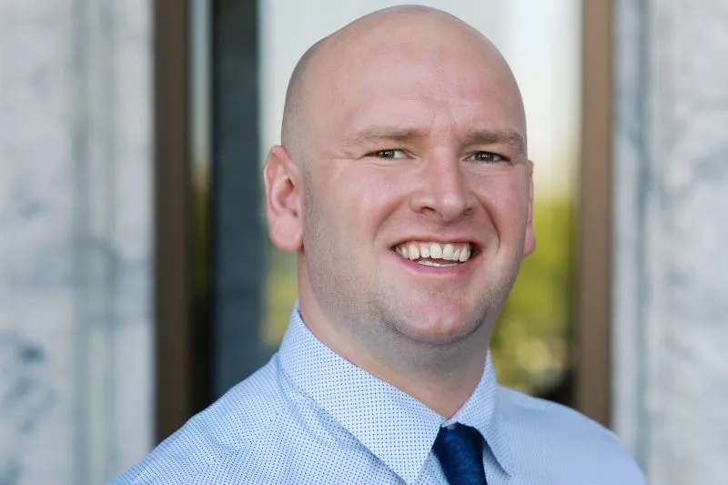 Professional headshot of a smiling bald man wearing a blue dress shirt and dark blue tie.