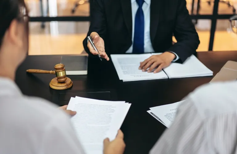 Two individuals in formal attire reviewing documents with a suited professional at a desk, accompanied by a wooden gavel.