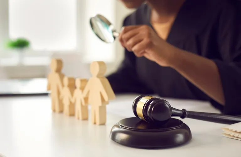 Person holding magnifying glass examining wooden family figures with judge's gavel in foreground.