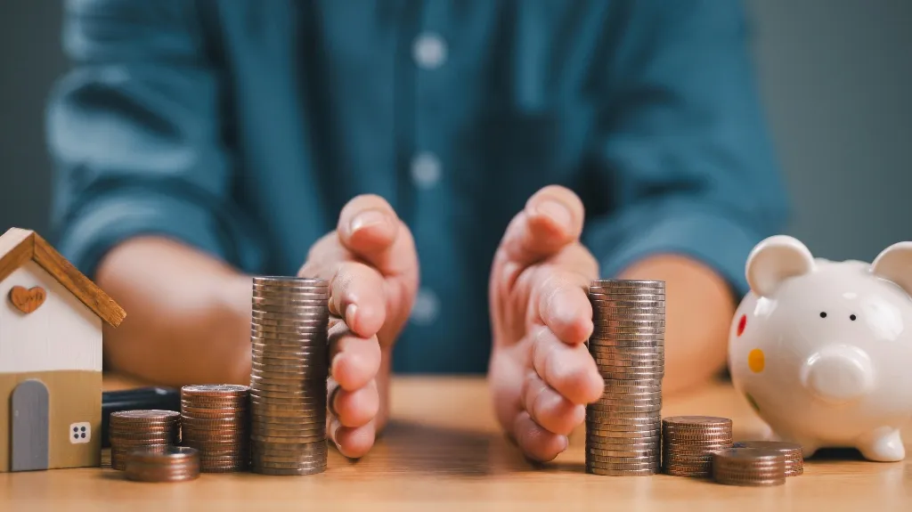 Hands protecting stacks of coins on a wooden table with a piggy bank and small house model.