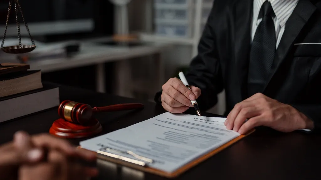Person in dark suit signing legal document on clipboard with gavel and scales nearby.