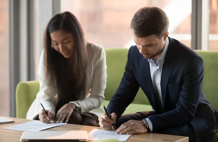 Two professionally dressed individuals, a woman and a man, seated on a green sofa, signing documents on a wooden table.