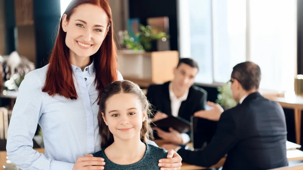 Smiling woman with red hair and young girl in foreground, two men in suits conversing in background.