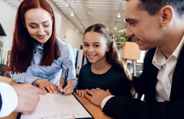 A woman signing a document at a table with a smiling young girl and a man in a suit.