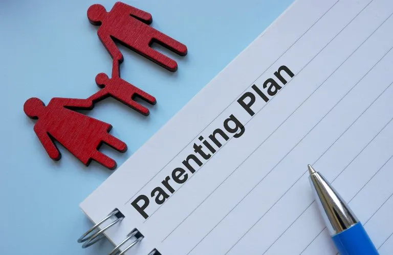 Spiral notebook with "Parenting Plan" title, blue pen, and red family figurines on light blue background.