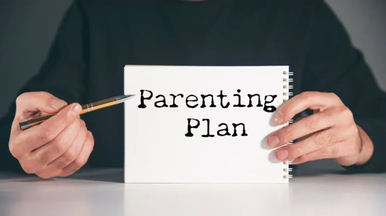Person in black shirt holding a white spiral notebook with text "Parenting Plan" and pointing with a pen.