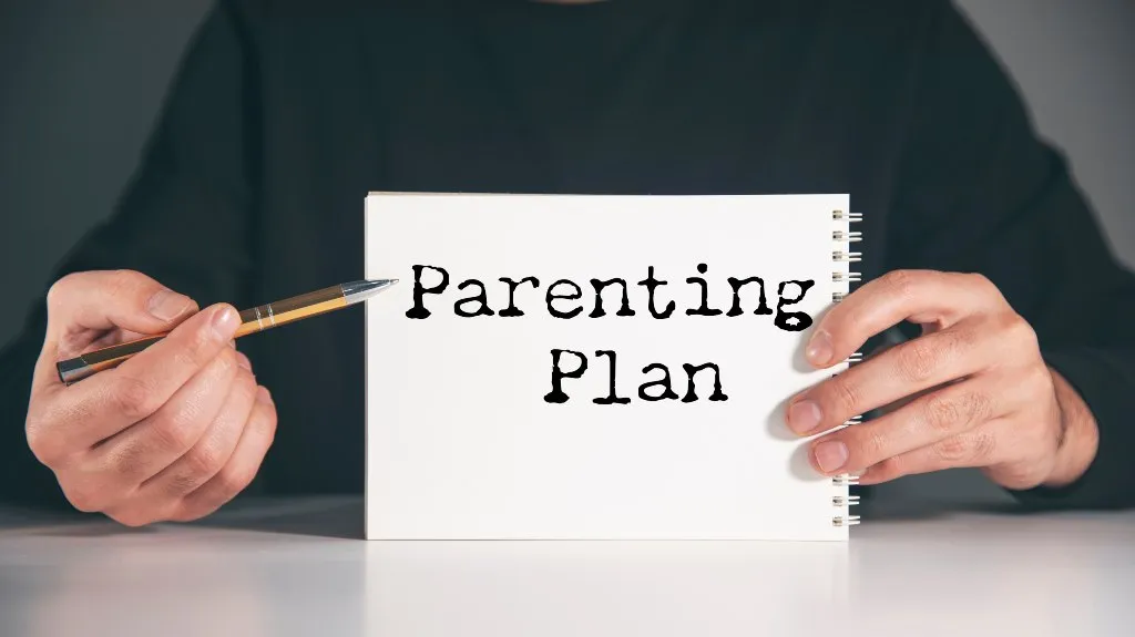 Person in black shirt holding a white spiral notebook with text "Parenting Plan" and pointing with a pen.