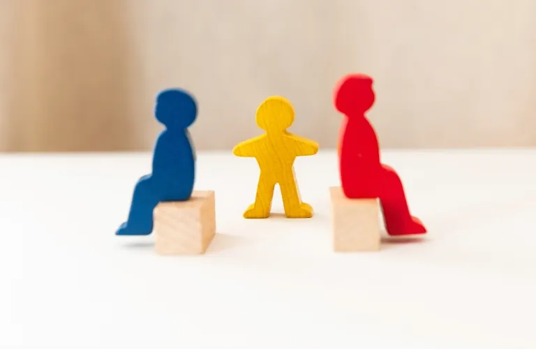 Three wooden figurines: a blue and a red seated figure on blocks, and a yellow standing figure between them.