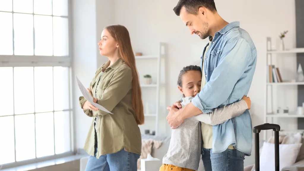 A man in a denim shirt hugging a child while a woman stands by a window holding papers.