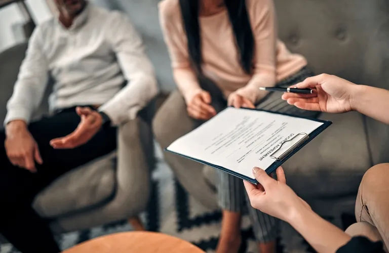 Three individuals seated on gray armchairs, one holding a clipboard with a document titled "CONSENT AGREEMENT.