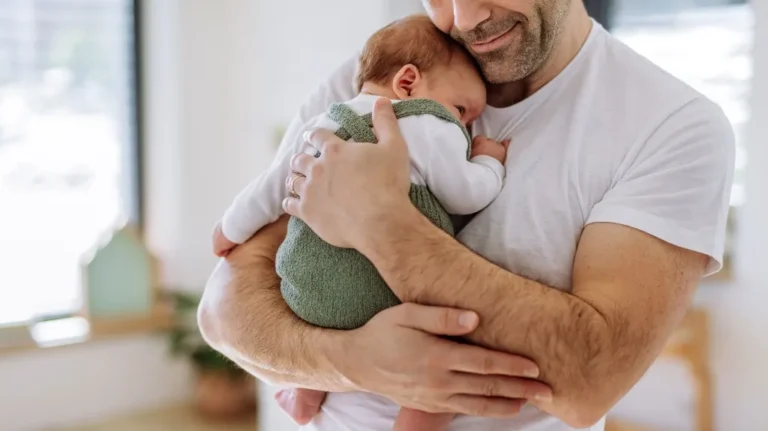 Man in white t-shirt gently holding and comforting a baby in green outfit indoors.