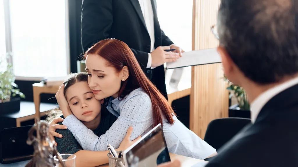 Woman with red hair comforting a young girl in an office setting with two men in suits.