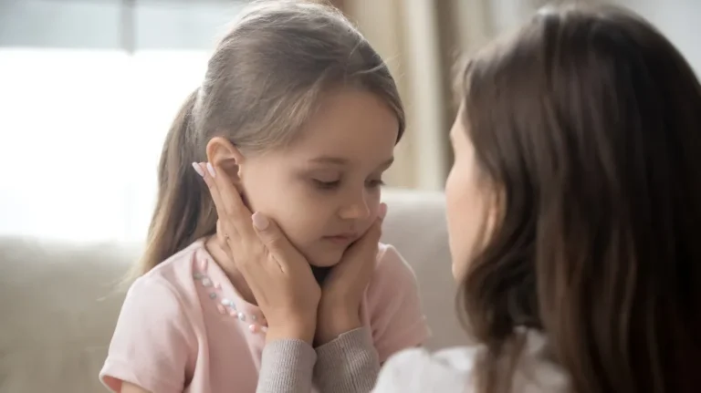 Adult woman gently holding young girl's face, both with brown hair, in a softly lit room.